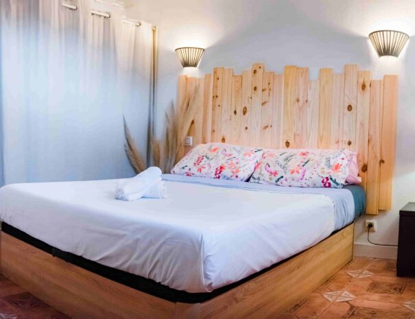 habitación doble muy luminosa con sabanas blancas y de flores rosas, con cabezal de madera y aplics en la pared