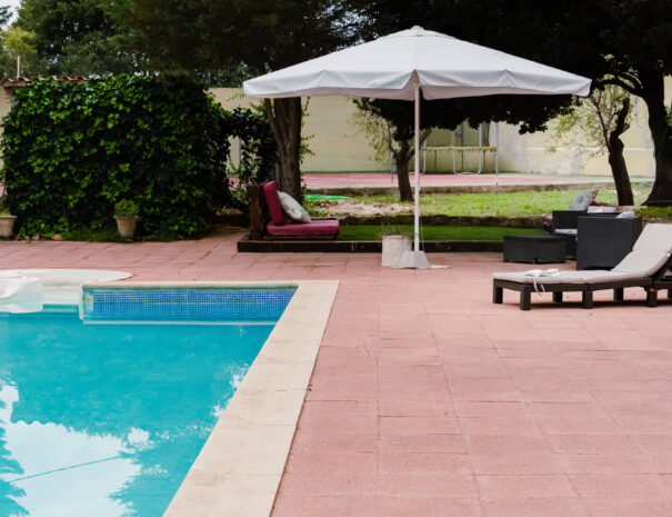 piscina azul con habaca, sillon, sombrilla blanca, zona chill out con cesped y psta de tennis con colchoneta en el fondo