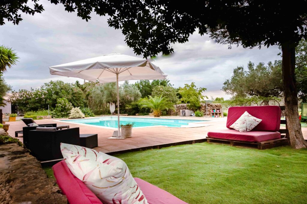 zona de jardín con cómodos sofás de pallets y cojines de flores, con piscina, butacas y sombrilla blanca