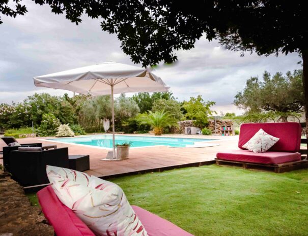 zona de jardín con cómodos sofás de pallets y cojines de flores, con piscina, butacas y sombrilla blanca